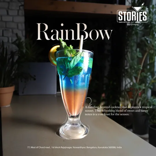 Blue-to-orange layered Rainbow cocktail with mint and an orange slice beside potted plants – drink poster for Stories Bar & Kitchen, Rajajinagar